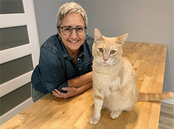 Lisa Grivois Leaning on Wooden Table with Orange Cat Sitting On Top